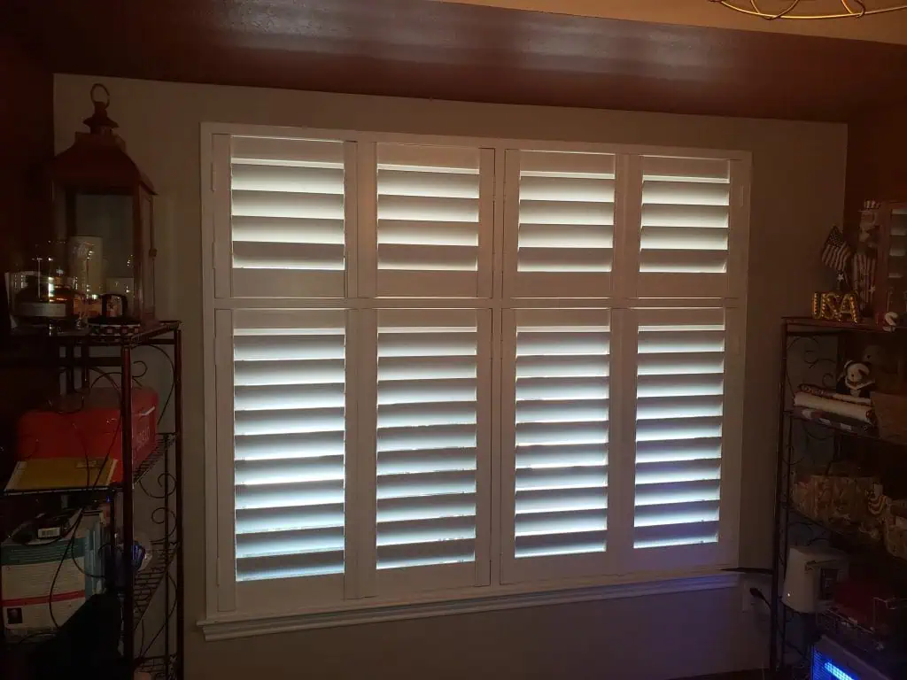 White plantation shutters elegantly cover a rectangular window. The slats are partially open, allowing light to gently filter through. Dark walls frame the window, complemented by shelves adorned with various decorative items.
