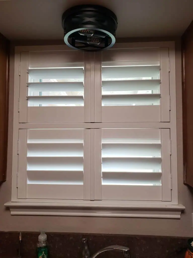 White plantation shutters drape elegantly over the window, allowing light to filter through their slats. Above, a sleek circular ceiling fixture illuminates the space, while below, a kitchen sink with a soap dispenser rests on the warm brown countertop.