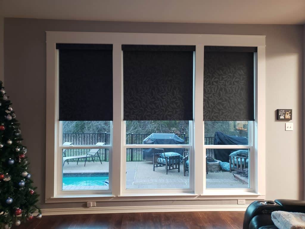 Large windows with dark roller shades partially drawn reveal an outdoor patio with chairs, a pool, and a barbecue grill. A Christmas tree is visible to the left, and some wall decor is on the right.