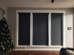 A room with three large, dark, patterned roller shades drawn shut. To the left stands a decorated Christmas tree adorned with ornaments. The wooden floor gleams, and a light switch is visible on the wall to the right.