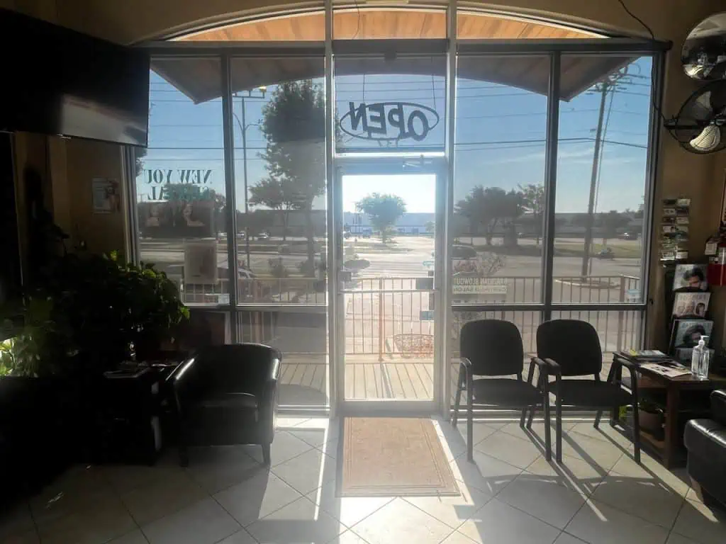 The interior of a sunlit waiting room features black chairs and a plant. A large glass door with an "OPEN" sign, flanked by stylish plantation shutters, leads outside. Posters adorn the windows, offering glimpses of the street and trees through the door.