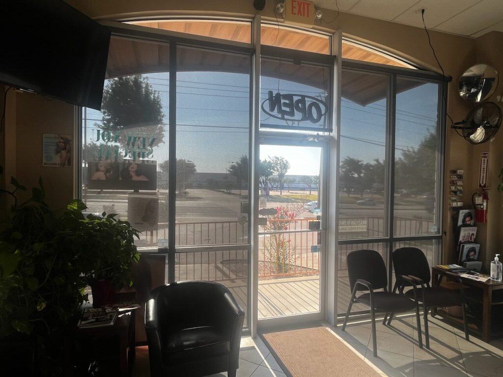 A waiting room features chairs lined up against a windowed wall. Sunlight softly filters through plantation shutters and glass doors with an "OPEN" sign. Outside, trees and a parking area are visible, while potted plants and wall decorations enhance the interior's charm.