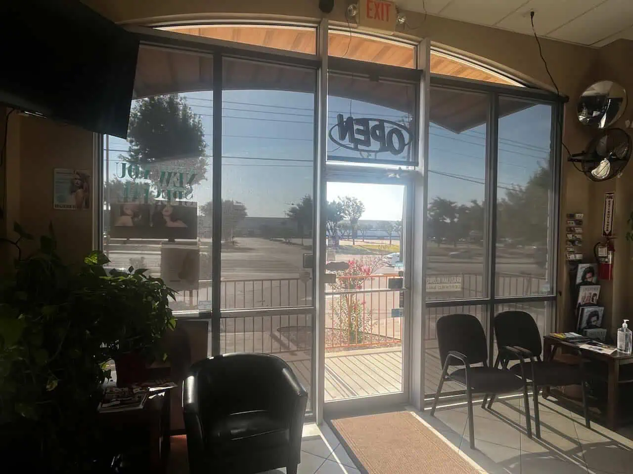 A waiting room features chairs lined up against a windowed wall. Sunlight softly filters through plantation shutters and glass doors with an "OPEN" sign. Outside, trees and a parking area are visible, while potted plants and wall decorations enhance the interior's charm.