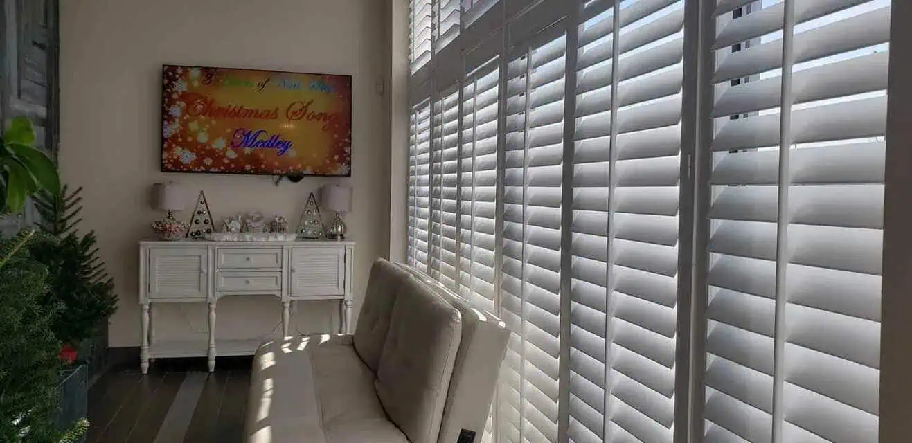 A sunlit room with white plantation shutters from Tarrant County covering tall windows. A beige couch is placed against the windows in front of a white console table decorated with small Christmas-themed ornaments and a TV mounted above, displaying a festive screen.