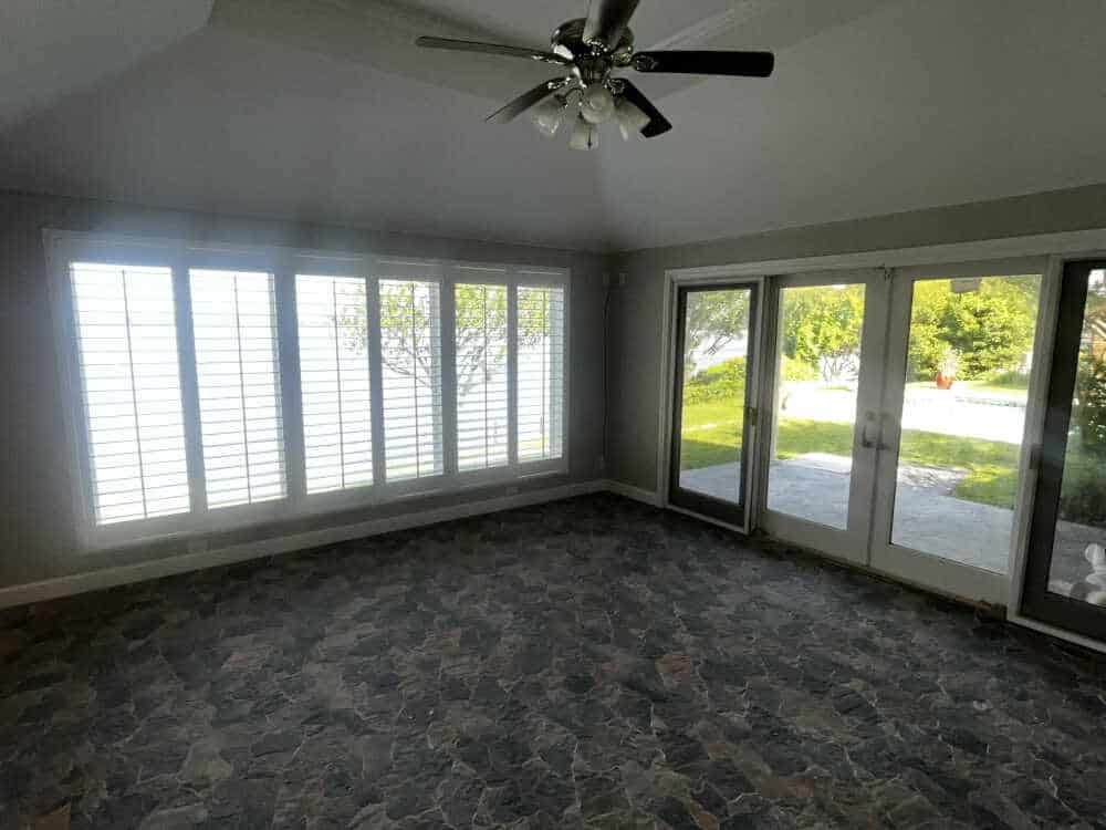 A room with large windows and french doors overlooking a garden. The windows have white plantation shutters, showcasing expert Shutter Installation in Tarrant County, and the floor is covered with dark, patterned tiles. The ceiling features a ceiling fan with light fixtures. Natural light brightens the space.