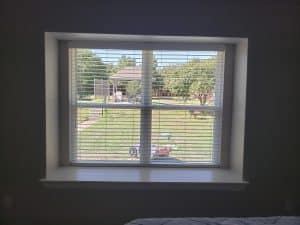 A window with white blinds partially opened reveals a sunny backyard with a pathway, vibrant green grass, and trees leading to a glimpse of another house. Shadows from the blinds gracefully fall across the interior wall.