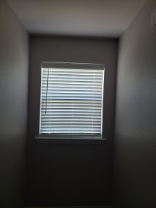 A small window with white blinds, partially open to let in light, is set in a plain, light gray wall between two narrow side walls.