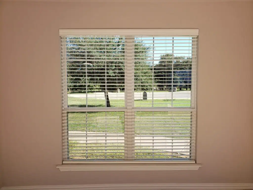 Partially open, the white blinds gently sway by the window, revealing a serene view of a grassy area with trees outside. Sunlight streams through, casting faint shadows on the beige walls inside.