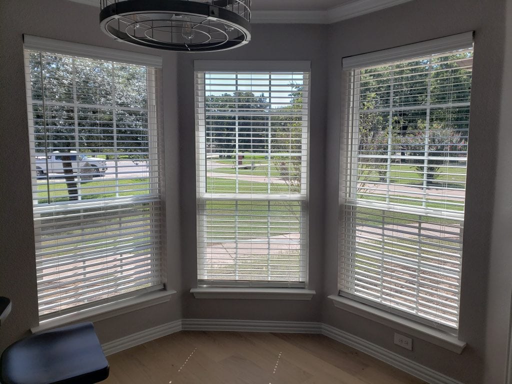 A sunlit room with three large windows, their white blinds elegantly filtering the light. The view reveals lush green grass, trees, and a quiet street. A circular light fixture graces the ceiling, while a dark chair peeks in from the left.