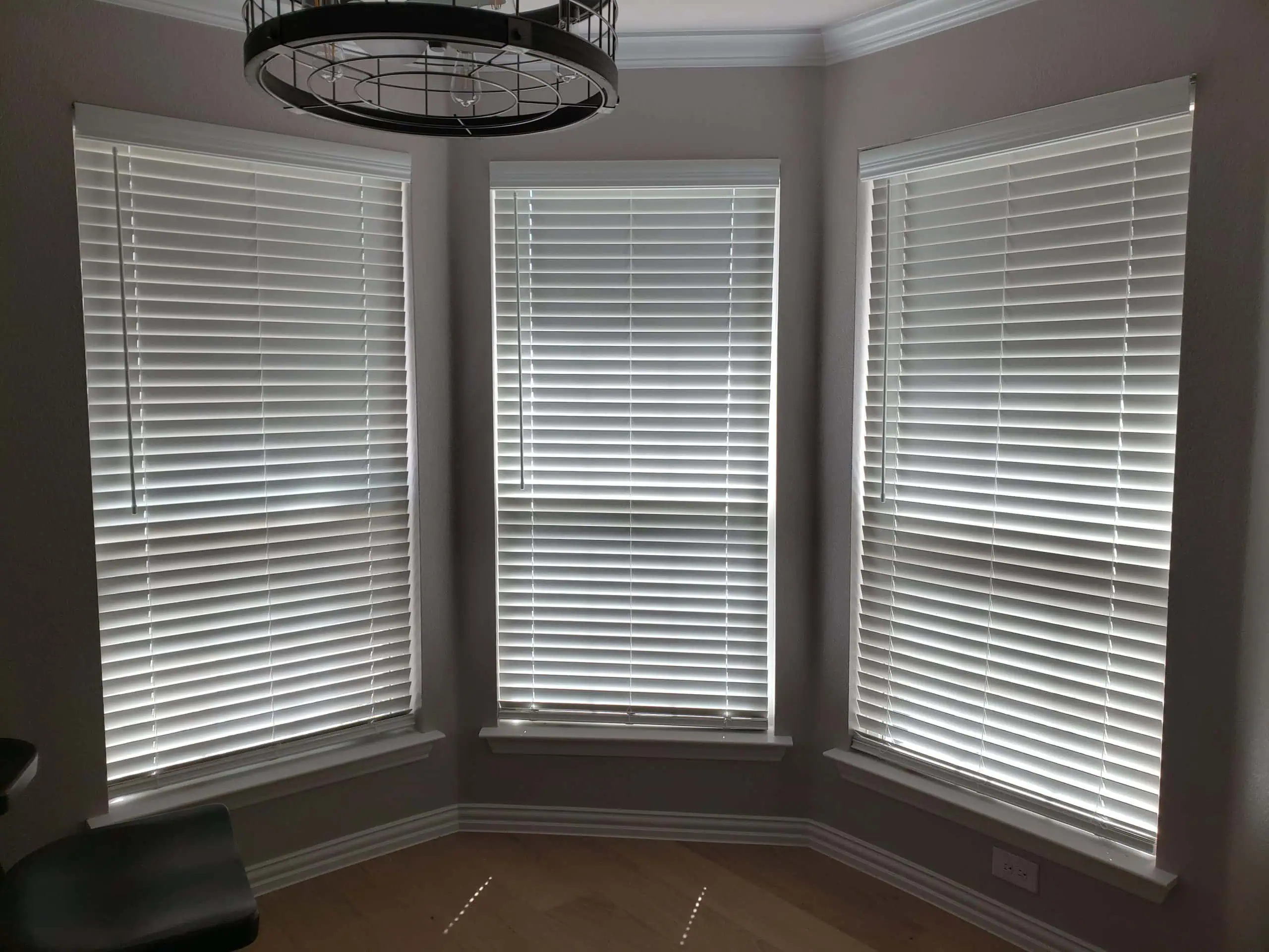 Three adjacent windows with pristine white blinds, neatly closed, adorn a softly lit room with cream walls and light wooden flooring. A modern circular chandelier elegantly hangs from the ceiling.