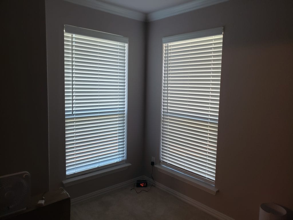 A room corner features two large windows with closed blinds, softly filtering light into the space. A digital clock on the floor displays red-lit numbers, while part of a fan sneaks into view on the left. The walls embrace a soothing light gray hue.