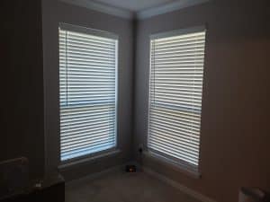 A room corner features two large windows with closed blinds, softly filtering light into the space. A digital clock on the floor displays red-lit numbers, while part of a fan sneaks into view on the left. The walls embrace a soothing light gray hue.