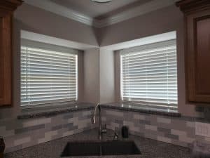 A kitchen sink with a granite countertop sits between two corner windows adorned with sleek white blinds. The backsplash boasts a gray and white tile pattern, complemented by wooden cabinets on either side.