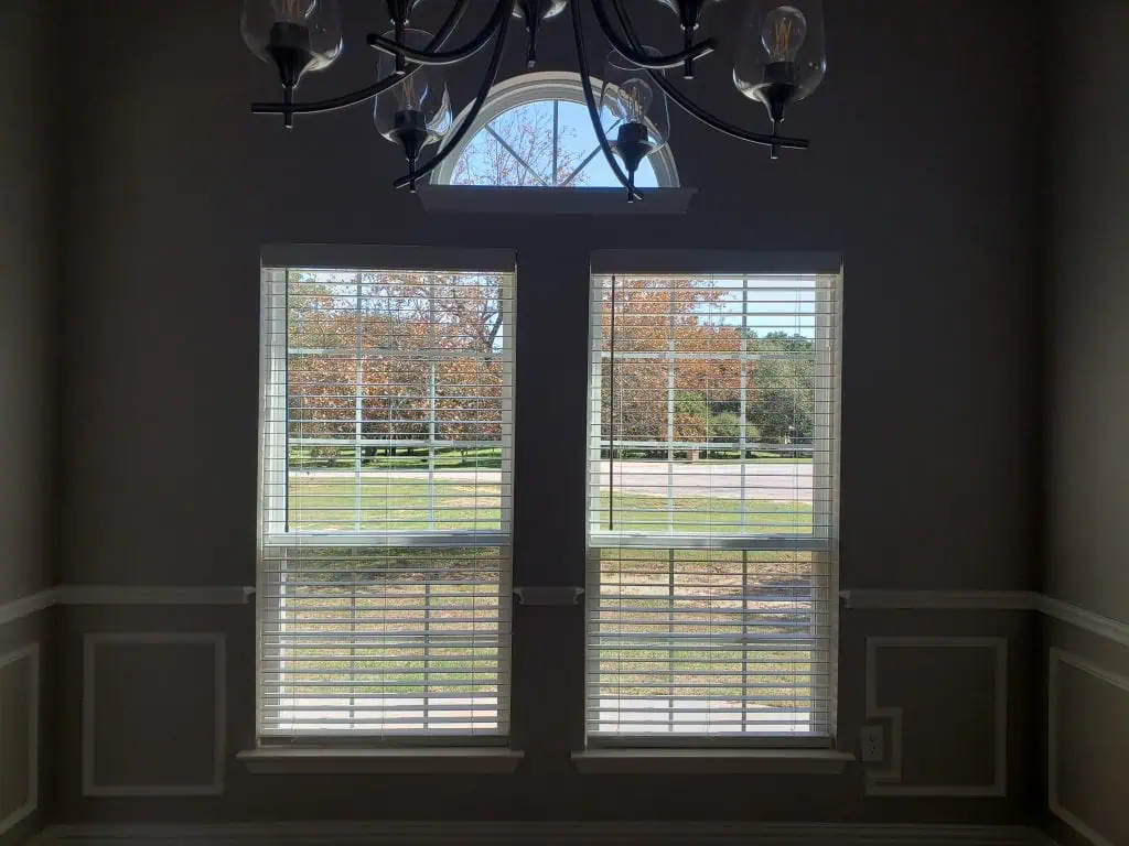 A dimly lit room with two tall, narrow windows featuring sleek white blinds. Outside, autumn trees flaunt their vibrant orange leaves. Above the windows is an elegant arched design with a chandelier gracefully suspended from the ceiling.