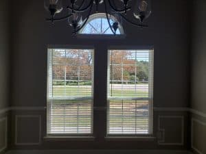 A dimly lit room with two tall, narrow windows featuring sleek white blinds. Outside, autumn trees flaunt their vibrant orange leaves. Above the windows is an elegant arched design with a chandelier gracefully suspended from the ceiling.