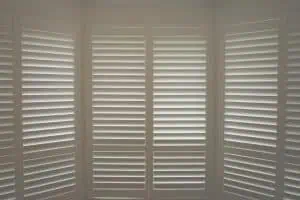 A room with large white plantation shutters Tarrant County partially closed, allowing slivers of sunlight to filter through, casting soft shadows inside. The shutters cover tall windows that appear to form part of a bay window setup. The overall ambiance is calm and serene.