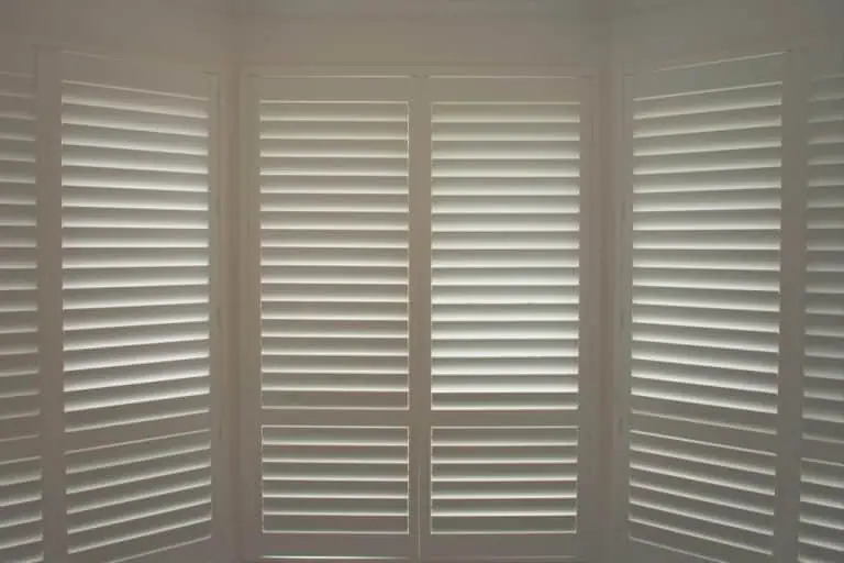 A room with large white plantation shutters Tarrant County partially closed, allowing slivers of sunlight to filter through, casting soft shadows inside. The shutters cover tall windows that appear to form part of a bay window setup. The overall ambiance is calm and serene.