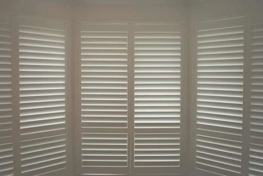 A room with large white plantation shutters Tarrant County partially closed, allowing slivers of sunlight to filter through, casting soft shadows inside. The shutters cover tall windows that appear to form part of a bay window setup. The overall ambiance is calm and serene.