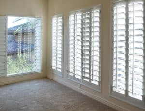 A bright room with large windows covered by white plantation shutters Tarrant County. The windows let in natural light, illuminating the beige carpet. Through the windows, palm tree fronds and neighboring buildings are visible. The room appears empty and spacious.