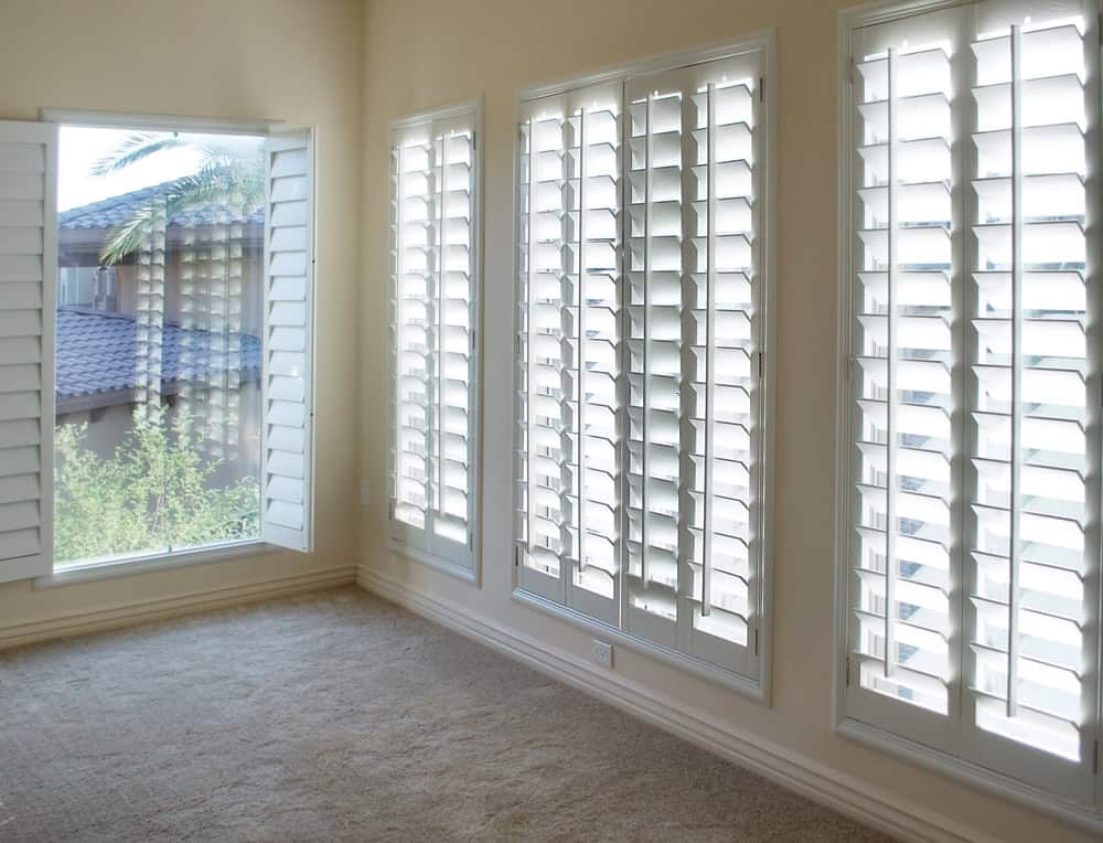 A bright room with beige walls and carpeted floors, featuring large windows adorned with white plantation shutters. Natural light filters through the partially open shutters, giving the room an airy atmosphere. A view of trees and a neighboring house is visible outside, showcasing expert shutter installation in Tarrant County.