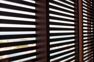 Through dark brown horizontal wooden blinds, a distant, blurred beach and ocean scene with clear blue water, sandy shores, and a few scattered buildings come into view. Light filters through the slats of these expertly crafted plantation shutters from Tarrant County, creating a pattern of shadows on the blinds.
