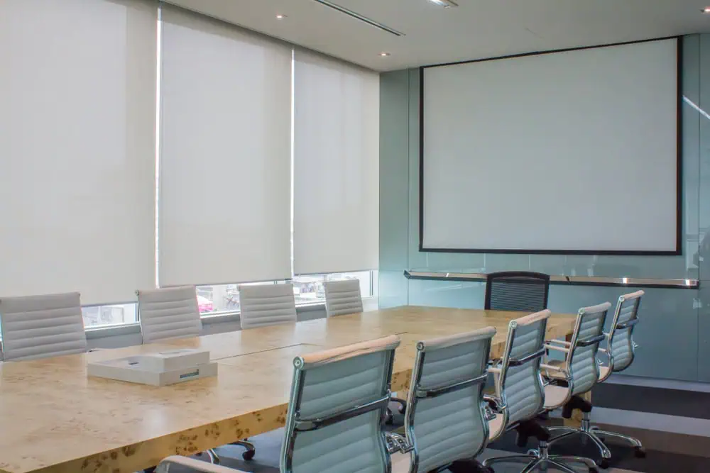 A modern conference room with a long wooden table surrounded by white swivel chairs. A blank projector screen is mounted on one wall, and large windows with Plantation Shutters Tarrant County cover the adjacent walls, allowing some natural light to filter in. Office supplies rest on the table.