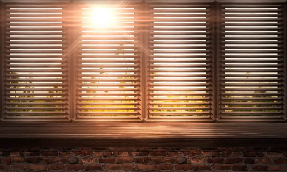 The image shows the sun shining through plantation shutters, casting a warm glow. The slats, part of a rustic brick wall structure beneath, highlight the texture of both the shutters and the bricks. This scene captures a cozy, serene atmosphere evocative of Shutter Installation in Tarrant County.