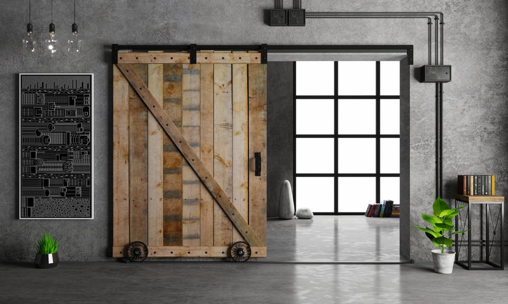 A stylish industrial-themed room features a large sliding wooden barn door with metal hardware from Barn Doors Tarrant County. On one side is a black and white artwork, a plant, and a small metal bookshelf with books. The background reveals a spacious, bright area with floor-to-ceiling windows.