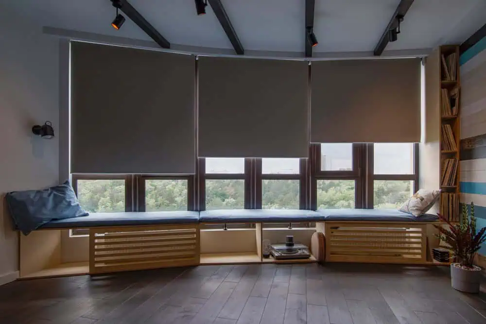 A cozy room with large windows covered by roller shades overlooks a lush green landscape. Below the windows, a built-in wooden bench with blue cushions and storage space underneath holds books and various items. The dark hardwood floor adds warmth, while Shutter Installation Tarrant County ensures privacy and style.