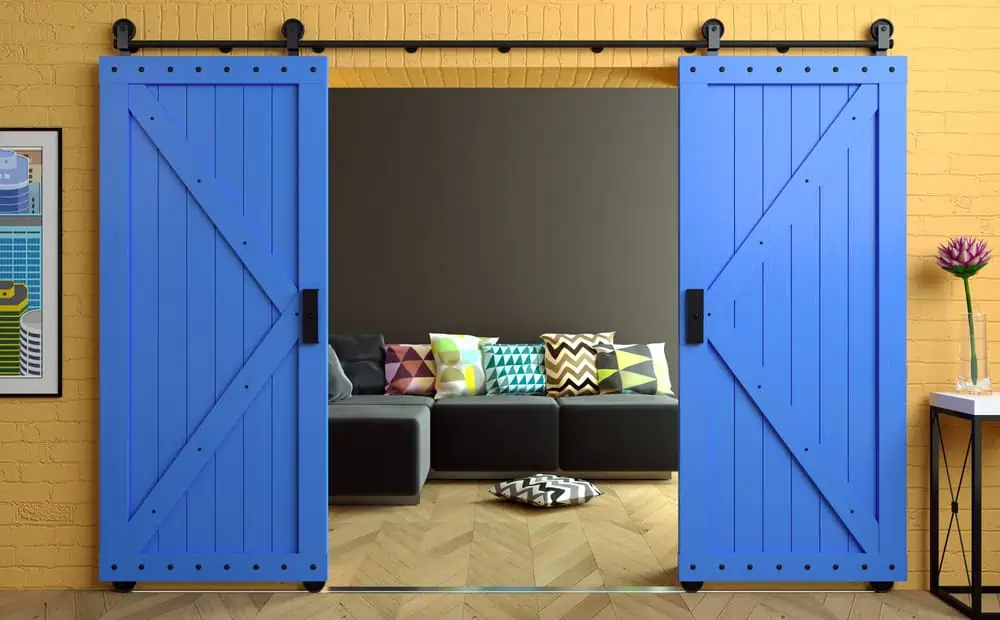 A modern living room with a black sofa adorned with colorful geometric-patterned cushions, visible through open bright blue sliding barn doors. The walls are yellow, featuring a black shelf displaying decorations to the right and framed artwork to the left. Elegant plantation shutters by Shutter Installation Tarrant County complement the overall aesthetic.
