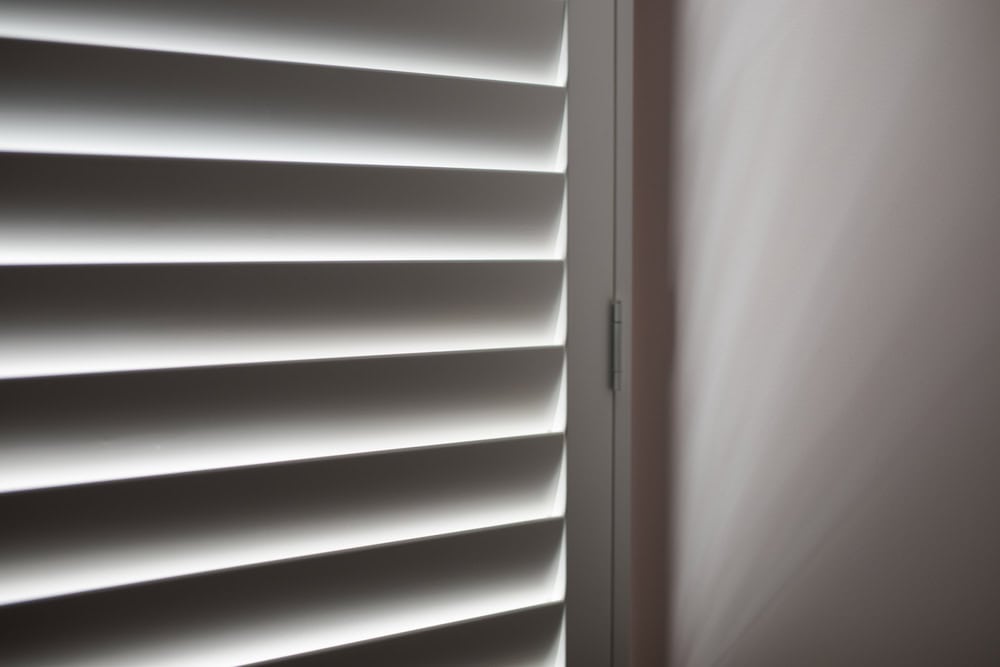 Close-up of partially opened window blinds with light streaming through, casting shadows on a nearby wall. The image creates a pattern of alternating light and dark stripes, giving a minimalist and modern feel—perfect inspiration for those considering blinds installation in Tarrant County.