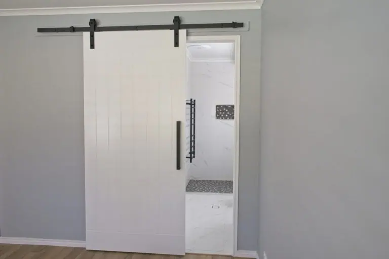 A minimalist room with light gray walls features a white sliding barn door, reminiscent of the Barn Doors Tarrant County offers, mounted on a black rail. The door is partially open, revealing a bathroom with white marble tiles and a sleek black towel rack.