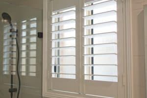 A close-up view of a bathroom with frosted glass windows covered by white plantation shutters. To the left, there is a glass shower door with a black showerhead visible inside. The scene is brightly lit by natural sunlight streaming through the windows, showcasing expert shutter installation in Tarrant County.