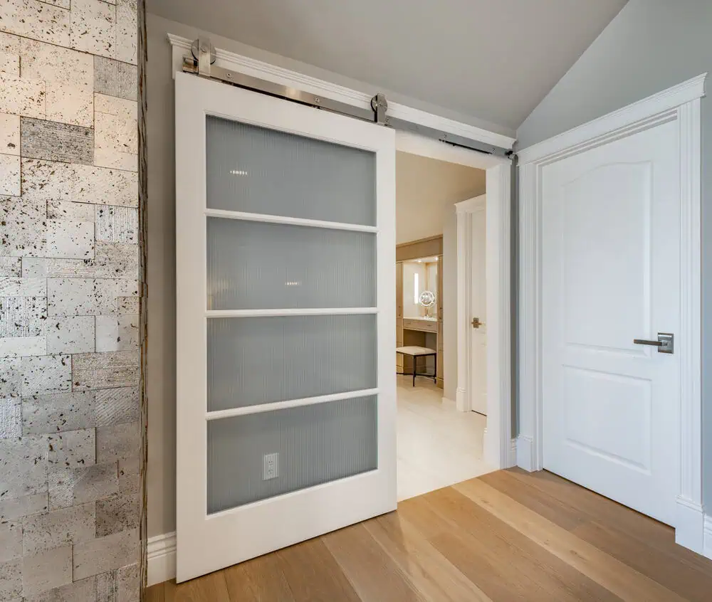 A white sliding barn door with frosted glass panels is partially open, revealing a room with beige walls and an alcove with a chair. The room has light wooden flooring, and an adjacent closed white door with a silver handle is visible to the right. For premium barn doors in Tarrant County, consider Barn Doors Tarrant County for top quality.