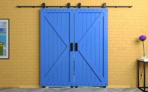 A pair of closed, bright blue barn-style sliding doors with black handles and hardware, mounted on a light yellow brick wall. Next to the door is a framed poster on the left and a small black table with a single purple flower in a white vase on the right, showcasing Shutter Installation Tarrant County.