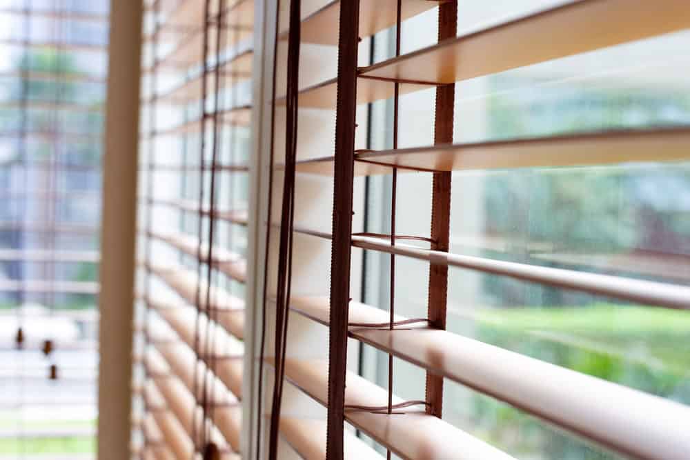 Close-up view of wooden window blinds with horizontal slats partially open, allowing light to filter through. The background shows a blurred outdoor scene with greenery, indicating a garden or yard outside. For expert blinds installation in Tarrant County, trust the professionals who understand quality and style.