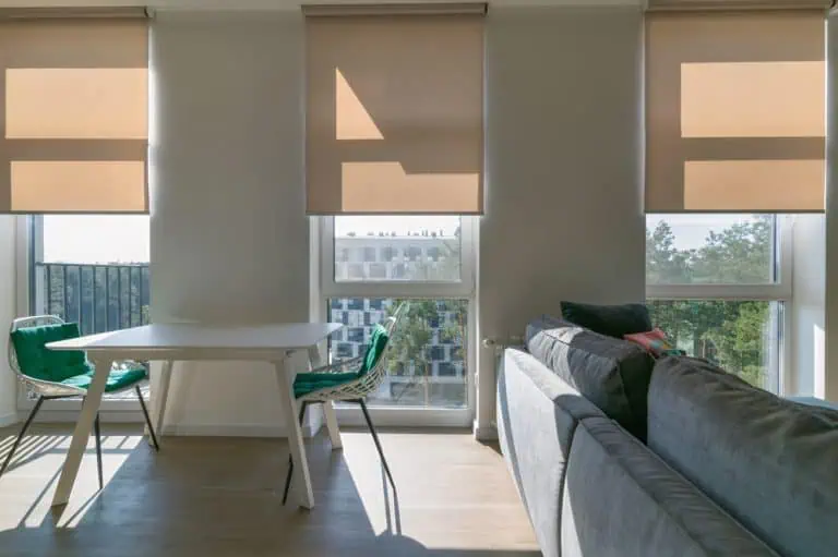 A modern, sunlit living space features a gray sofa on the right, a white dining table with two green chairs on the left, and three large windows with plantation shutters installed. An urban view outside includes a building with trees and a partially visible balcony railing.