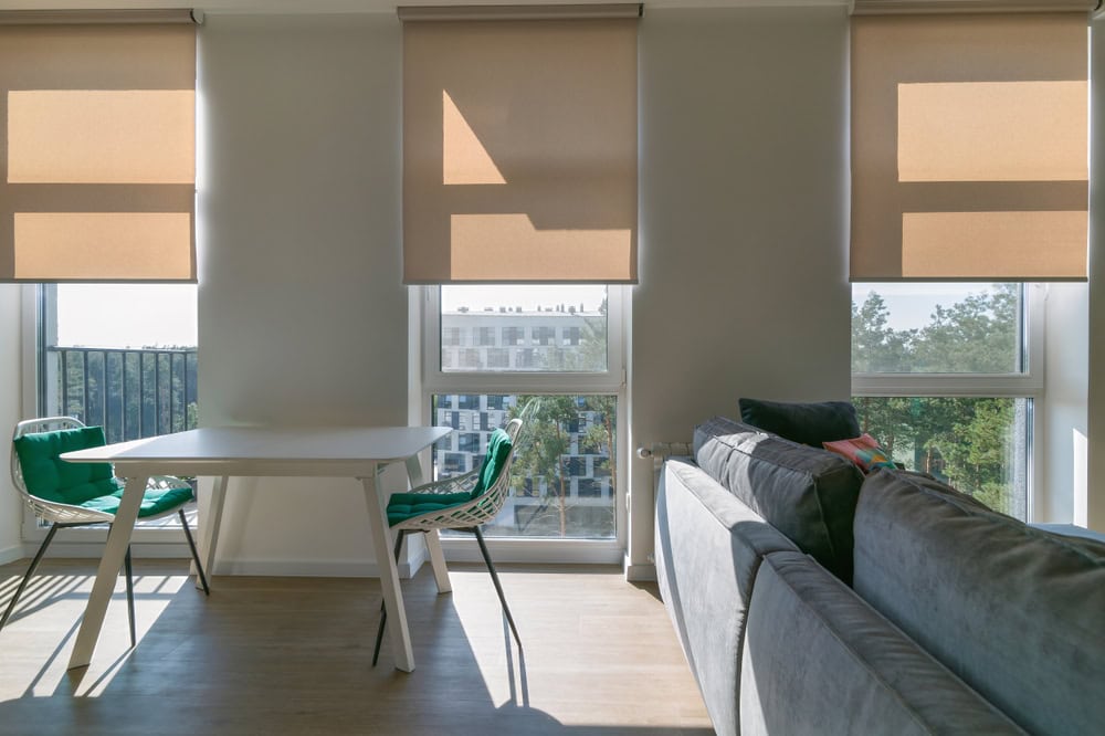 A modern, sunlit living space features a gray sofa on the right, a white dining table with two green chairs on the left, and three large windows with plantation shutters installed. An urban view outside includes a building with trees and a partially visible balcony railing.