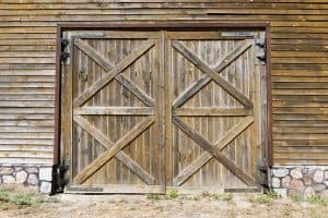 A large wooden barn door, constructed with vertical planks and an X-brace pattern, is prominently displayed. The door is framed by a wooden wall featuring horizontal siding, and the base is lined with a row of stones. Grass grows at the bottom edge, reflecting the rustic charm often seen in Barn Doors Tarrant County.
