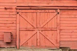 A rustic red barn door with a large X pattern made of wooden planks, weathered paint showing signs of aging. A small red mailbox adorns the lower left side of the barn wall. The ground in front is bare and slightly uneven, typical for Barn Doors Tarrant County specialties.