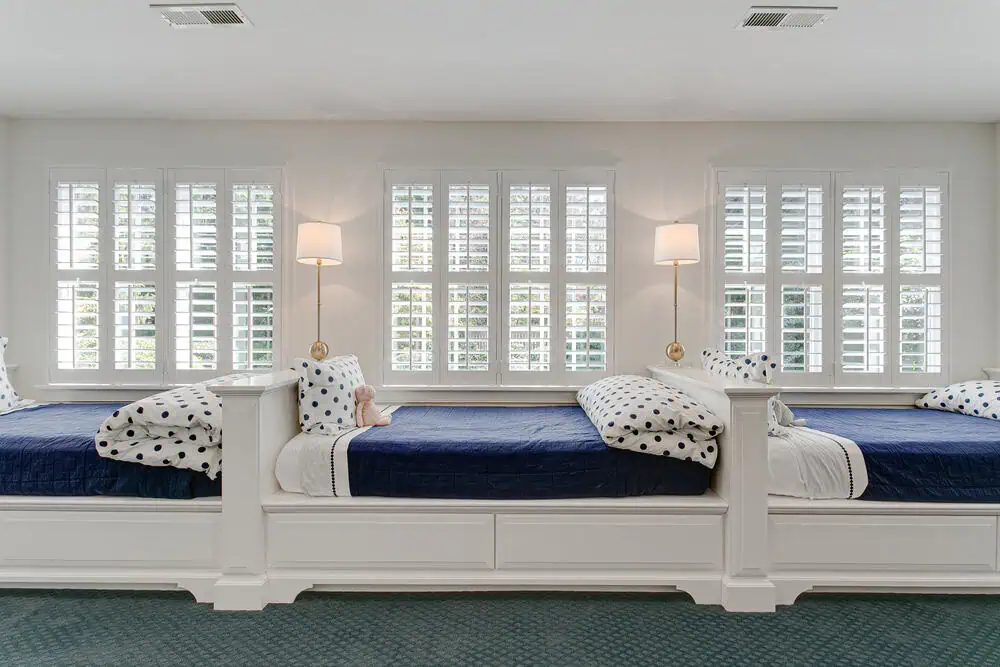 A bright room with three single beds aligned horizontally beneath large windows boasting Plantation Shutters Tarrant County residents would admire. Each bed has blue bedding and polka-dotted pillows, while two wall-mounted lamps with white shades are spaced between the beds, creating a symmetrical arrangement.