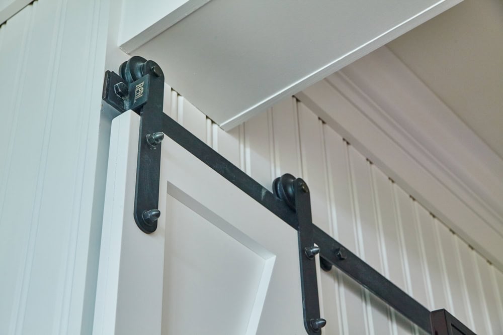 A close-up of the upper section of a white barn door with black sliding hardware, reminiscent of elegant Plantation Shutters Tarrant County. The door features vertical paneling, and the black metal rollers are attached to a track mounted on the door frame, allowing it to slide open and closed effortlessly.