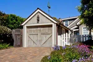 A single-car garage with weathered wooden doors sits beside a house with light siding and blue plantation shutters in Tarrant County. The driveway is paved with bricks, and the yard features blooming purple flowers, green plants, and a white picket fence under a clear blue sky.