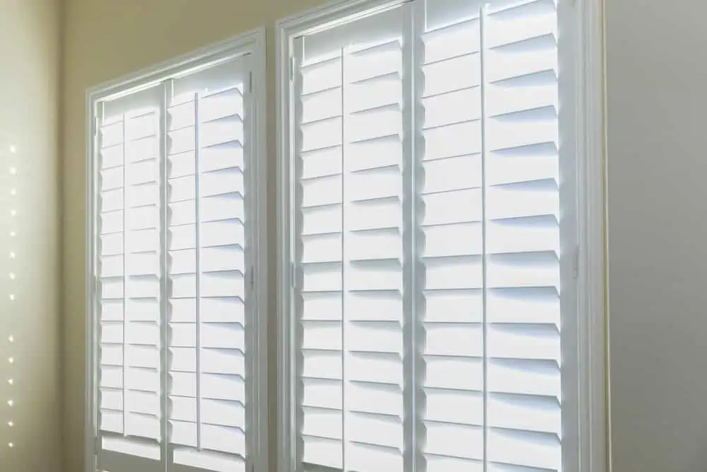 Two large windows covered with white plantation shutters. Sunlight filters through the shutters, creating a bright and airy atmosphere in the room. The slats are angled to allow light in while maintaining privacy. Installed by experts in shutter installation Tarrant County, the window frames are clean and modern.