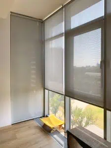 A bright room with large windows covered by light grey roller blinds installed by Blinds Installation Tarrant County. Sunlight filters in, illuminating the wooden floor. A small yellow chair is situated by the window, facing the outdoor view of a green lawn and trees.