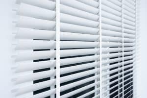 A close-up view of white horizontal window blinds partially open, allowing light to filter through the gaps. The blinds are evenly spaced and aligned, with a dark background seen between the slats. For a seamless look like this, consider professional shutter installation in Tarrant County.