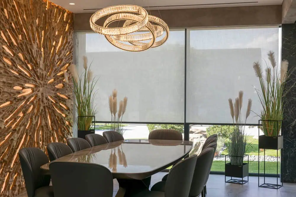 A modern dining room with a polished wooden table surrounded by eight upholstered chairs. Unique light fixtures hang above the table, and a decorative wall feature with starburst design is on the left. Tall plants in pots flank both sides of the large window, which boasts sleek plantation shutters from Tarrant County.