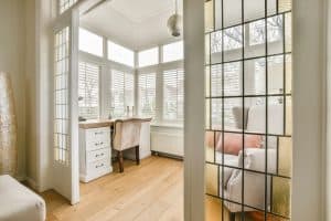 A cozy sunroom with large windows featuring Plantation Shutters Tarrant County. The room includes a desk with a white chair, a cushioned armchair with pink and gray pillows, and a wooden floor. Decorative glass panels adorn the entrance doors.