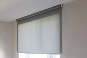 A gray roller shade is fully drawn down, covering a window set within a beige-colored wall. The minimalistic and neat design suggests a modern interior. The shade is mounted at the top of the window, with soft natural light filtering through—a perfect complement to Plantation Shutters Tarrant County installations.