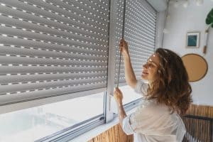 A woman with curly hair, wearing a white shirt, is seen adjusting the plantation shutters of a window. She is smiling and standing in a bright room with string lights, a round mirror, and a plant on the wall. Light filters through the shutters.